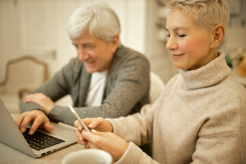 Joyful retired woman with short hairstyle relaxing at home with mobile phone, browsing newsfeed on social network, watching videos, her husband sitting in background, using laptop for distant work