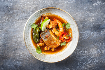 Modern style traditional Mexican seafood pozole soup with fish, king prawns and hominy in a clear sauce served as top view in design bowl