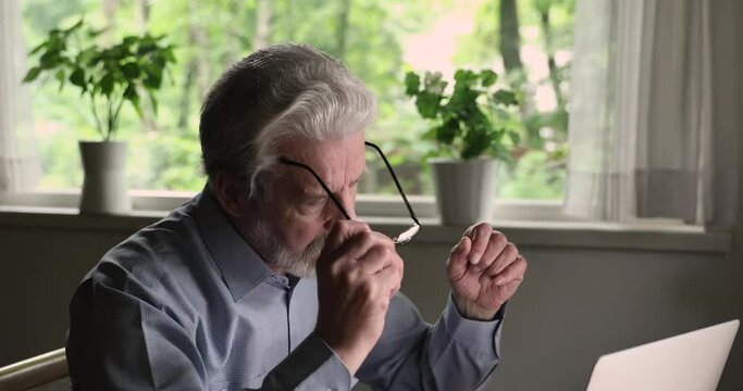 Head shot focused middle aged old man taking off glasses, making pause in computer work, suffering from dry eyes syndrome or relieving eyes strain, continue working online using modern tech gadget.