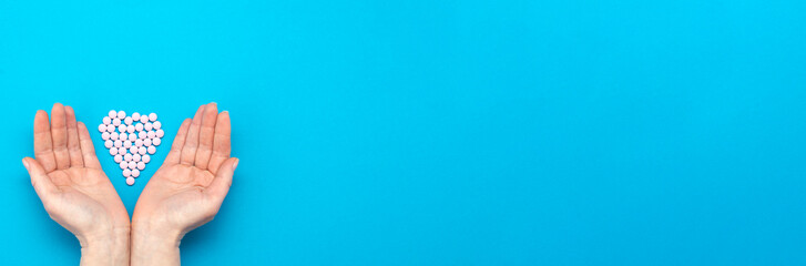 Pink pills in the shape of a heart on a blue background and female hands surround a heart made of pills