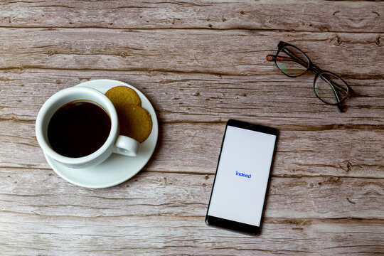 01-30-2021 Portsmouth, Hampshire, UK A Mobile phone or cell phone laid on a wooden table with indeed app opening also a coffee and glasses