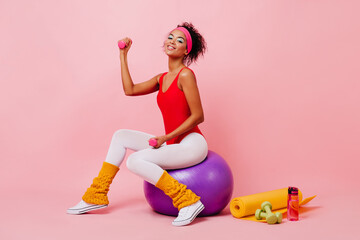 Stunning woman in bodysuit sitting on fitness ball. Sporty african girl posing with dumbbell on...