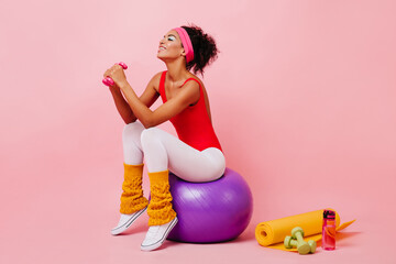 Smiling black girl with dumbbells sitting on pink background. Laughing african sporty woman training with fitness ball.