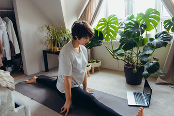 A woman on a mat trying to do a twine asana position in her bedroom with plants. Online class yoga...