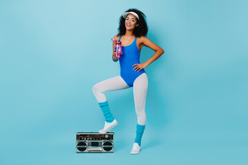 Attractive african lady in aerobics outfit standing on boombox. Studio shot of wonderful black girl drinking water after training.