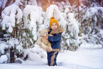 Cute baby boy playing with teddy bear outdoors. Kid having fun in winter snowy pine forest. Snow in frosty winter Park. Outdoors. Winter and Christmas time concept. Activity on fresh air concept.