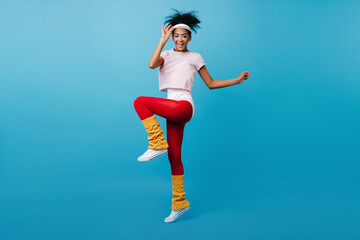 Inspired black woman enjoying aerobics. Full length view of glamorous african girl jumping on blue background.