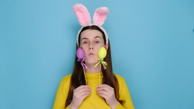 Cute comic young woman wears bunny ears makes funny face, plays fool, holds colored eggs, has happy expression, dressed in sweater, models over blue background with copy space. Easter holiday concept