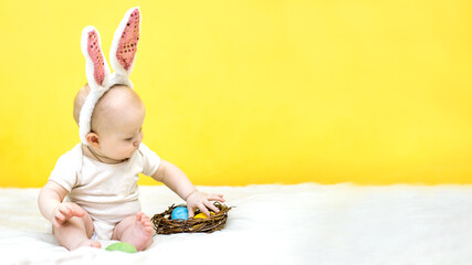 banner with child sitter with Easter bunny ears decorated with sequins sits near yellow wall. baby...