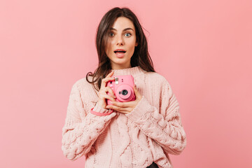 Gray-eyed lady with astonished expression makes a photo on a pink mini camera. Woman with straight dark hair in knitted sweater posing on an isolated background