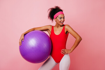 Carefree girl in red bodysuit doing aerobics. Studio shot of african female model with fitness ball...