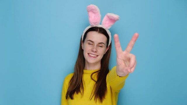 Portrait of attractive happy brunette woman in pink bunny fluffy ears is showing peace gesture, wears yellow sweater, models over blue studio background. Easter holiday and victory sign concept