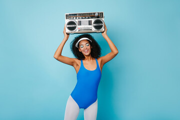 Amazing fitness girl enjoying music on blue background. Happy african young woman with boombox...