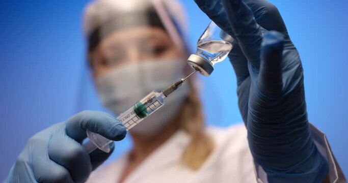 Female doctor wearing protective face shield and white uniform holding a syringe and preparing for covid 19 or other vaccine injection.