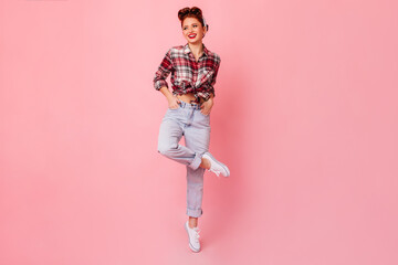 Full length view of ecstatic ginger woman standing on one leg. Studio shot of positive pinup girl in checkered shirt dancing on pink background.