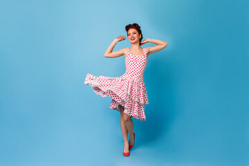 Glamorous ginger girl dancing on blue background. Studio shot of stunning pinup woman in polka-dot dress.