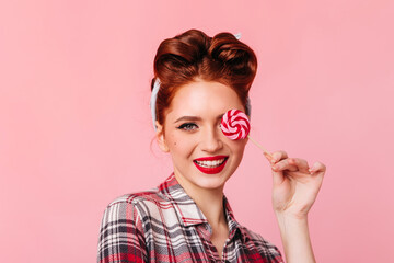 Smiling pinup girl eating red lollipop. Front view of woman in checkered shirt isolated on pink background.