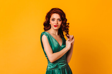 Spectacular young woman touching curly ginger hair on yellow background. Studio shot of amazing female model in green dress.