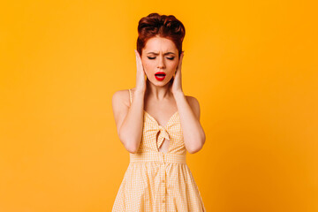 Upset pinup female model covering ears with hands. Studio shot of attractive caucasian woman isolated on yellow background.