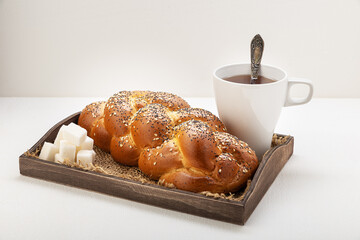 Wicker wheat bread sprinkled with sesame seeds and cereals on a wooden tray with a white mug with tea, a spoon, and lump sugar on a light background