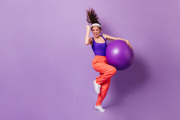 Woman in sports cap and bright top rejoices victory. Portrait of girl in red pants with fitball on purple background - obrazy, fototapety, plakaty