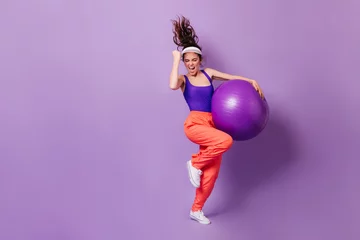 Fotobehang Woman in sports cap and bright top rejoices victory. Portrait of girl in red pants with fitball on purple background © Look!