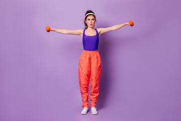 Full-length shot of girl in wide sweatpants and cap doing exercises for hands with dumbbells on lilac background