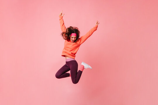 Lovely Brunette Girl In Pink Sports Headband And Tracksuit Jumps On Isolated Pink Background