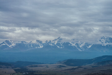 Scenic mountain landscape with great snowy mountain range among low clouds and green forest in valley at early morning. Atmospheric alpine scenery with blue white high mountain ridge under cloudy sky.