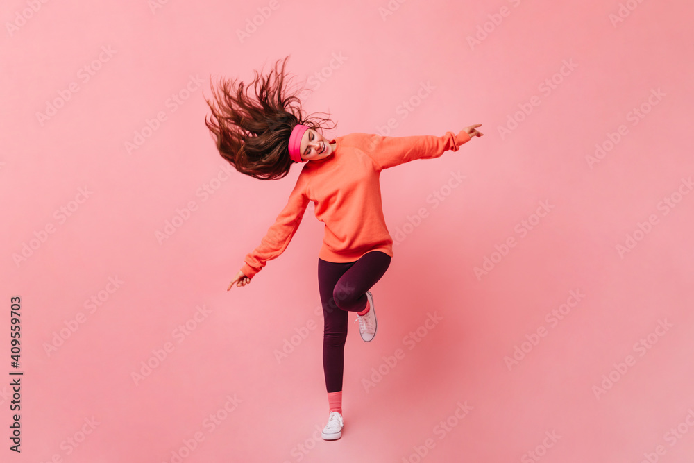 Poster Young lady in sports outfit dancing on pink background. Full length portrait of woman in orange sweatshirt