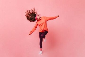 Young lady in sports outfit dancing on pink background. Full length portrait of woman in orange sweatshirt