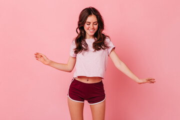 Cute brunette woman in summer outfit of sporty style is having fun dancing on pink background