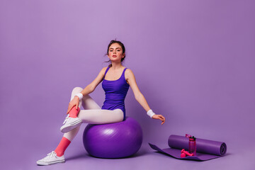 Young girl does aerobics on purple background. Woman sits on fit ball next to yoga mat
