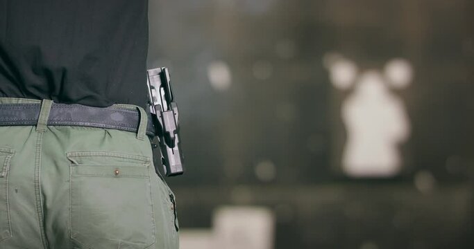 Back view close-up of a man pulling a pistol out of its holster and firing a shot at a shooting range.