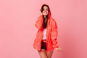 Cute dark-haired woman in white T-shirt and orange windbreaker laughs on pink background © Look!