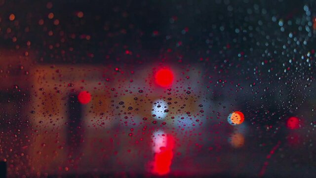 Raindrops run down the glass of the car against the backdrop of the bright lights of the night city. Rainy night. Cars pass the intersection. Bright colored traffic lights.