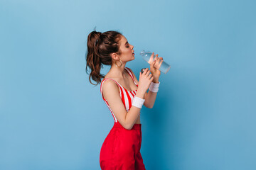 Dark-haired girl with massive earrings in red top and trousers drinking water on blue background