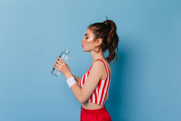 Young girl with highlighter on face drinks water. Portrait of woman with ponytail on blue background