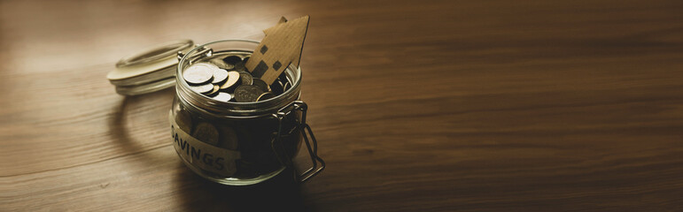 Selective focus of jar full of coins with tag saving and house model on it on wooden background with copy space.