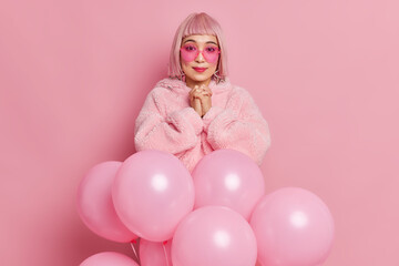 Fototapeta na wymiar Pretty glamour woman has hopeful expression keeps hands together wears trendy sunglasses winter fur coat poses in front of helium balloons against pink background going to celebrate festive event