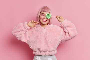Fashionable cheerful girl with bob hairstyle wears pink hair wig covers eye with green candy on stick wears fur coat poses indoor against rosy background. Asian young female model holds lollipop