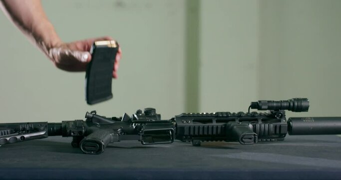 Close-up of a man examining and picking up a gun magazine clip and an assault rifle from a table.