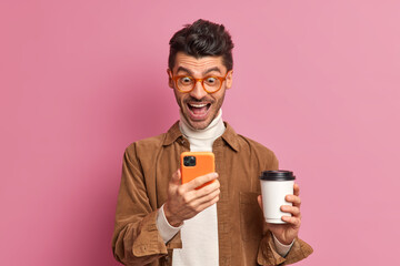 Cheerful man looks gladfully at smartphone screen reads funny news drinks takeaway coffee wears optical glasses brown shirt poses indoor against pink background. People emotions technology concept