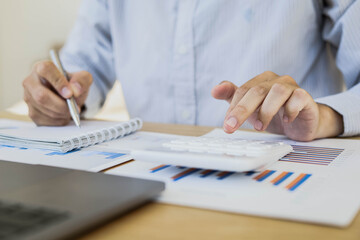 A business man is pressing a calculator to calculate sales in a sales graph data sheet. He's sitting in his private office in the company. Sales management concept.