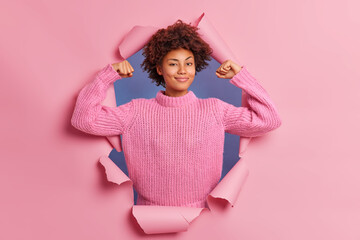 Self assured beautiful young Afro American woman raises arms shows biceps being strong and powerful proud of her own achievements wears casual knitted sweater breaks through paper background - obrazy, fototapety, plakaty