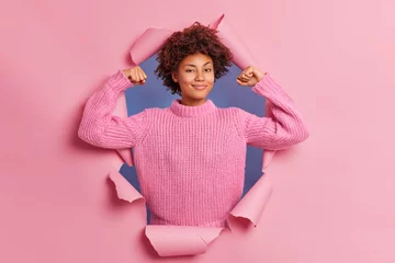 Foto op Canvas Self assured beautiful young Afro American woman raises arms shows biceps being strong and powerful proud of her own achievements wears casual knitted sweater breaks through paper background © Wayhome Studio