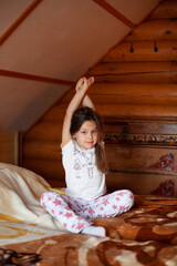 a charming, sleepy girl is sitting in her pajamas on a sunny morning on the bed in the bedroom of a rustic log house and sipping her hands on exercise.