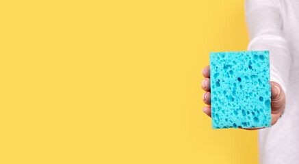 Hand with turquoise cleaning sponge on yellow background.