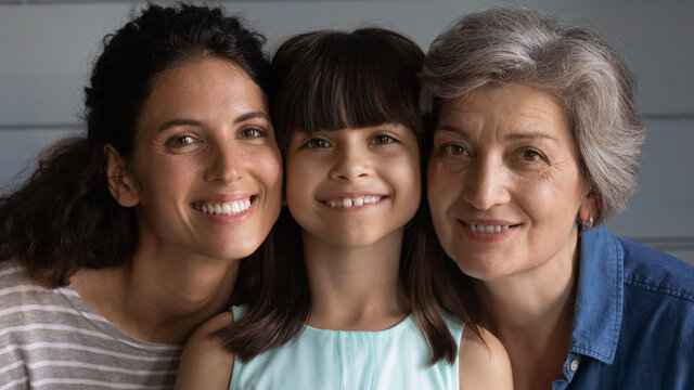 Head shot portrait close up smiling little girl with mature grandmother and young mother looking at camera, three generations of women touching cheeks, posing for photo, growing process concept