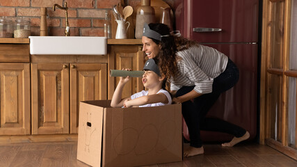 Overjoyed mother with little daughter playing pirates in kitchen, wearing handmade costumes, happy mum pushing cardboard box ship with cute girl inside, holding tube as telescope, having fun